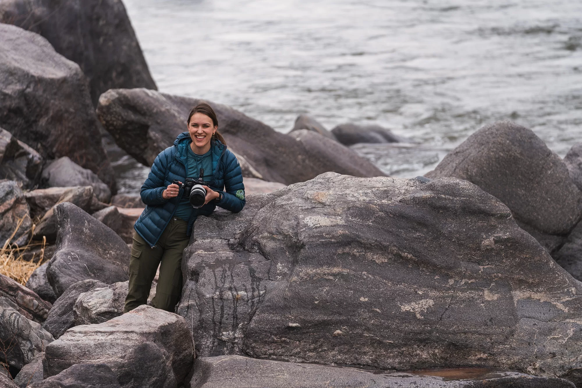 A photographer leans against a rock next to a river and smiles at the camera.