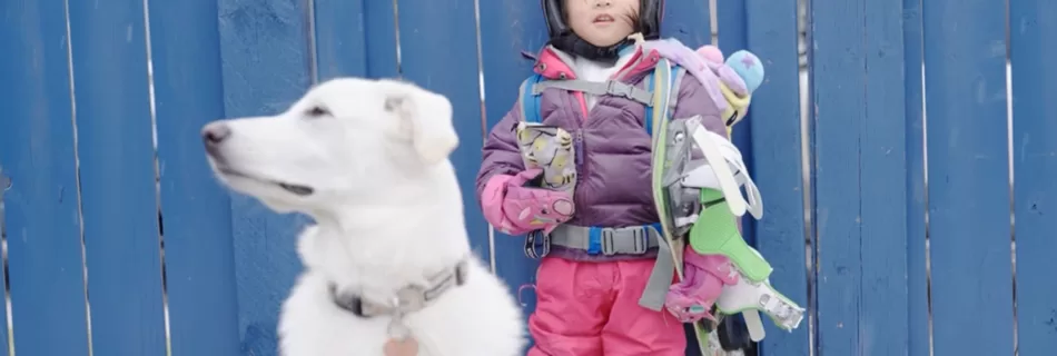 A young girl dressed in colorful snowboarding gear stands against a blue fence with her fluffy white dog sitting in the foreground.
